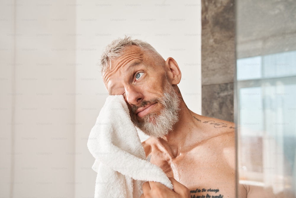 Vista verticale dell'uomo barbuto maturo che si pulisce il viso con un asciugamano bianco al mattino. Maschio che ha il concetto di routine di bellezza quotidiana