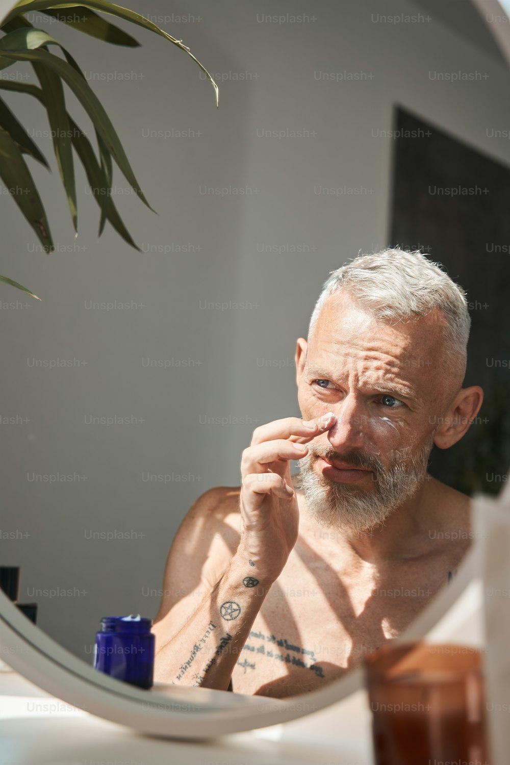 Reifer Mann, der kosmetische Creme auf sein Gesicht aufträgt, während er den Spiegel im Badezimmer betrachtet. Konzept der Gesichtspflege und -hygiene. Häuslicher Lebensstil. Grauhaariger europäischer Rentner mit Tattoos