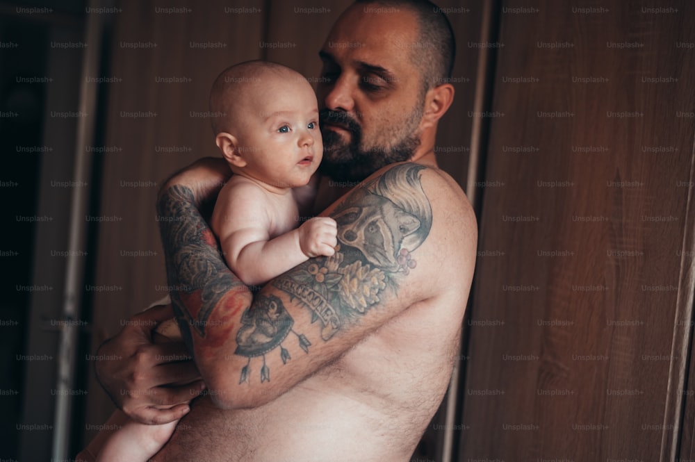 Padre barbudo tatuado jugando con su hijo en pañales mientras está en casa