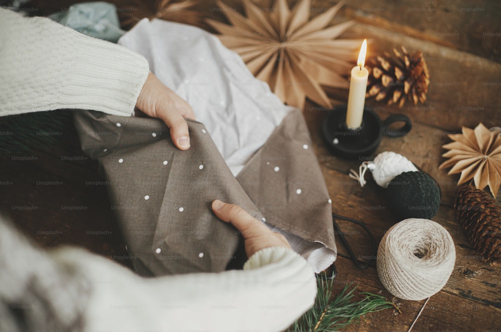 Furoshiki Christmas gift. Hands wrapping christmas gift in brown fabric on rustic wooden table with scissors, craft paper star, candle. Atmospheric moody time, nordic style. Zero waste holiday