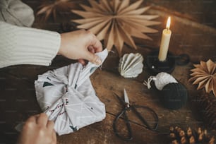 Furoshiki Christmas gift. Hands wrapping christmas gift in modern fabric on rustic wooden table with scissors, craft paper star, candle. Atmospheric moody time, nordic style. Zero waste holiday