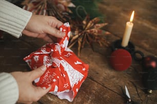ハサミ、紙の星、ろうそく、装飾品を備えた素朴な木製のテーブルの上に赤いお祝いの布でクリスマスプレゼントを包む手。雰囲気のあるムーディーな時間。ゼロウェイストホリデー、風呂敷クリスマスプレゼント