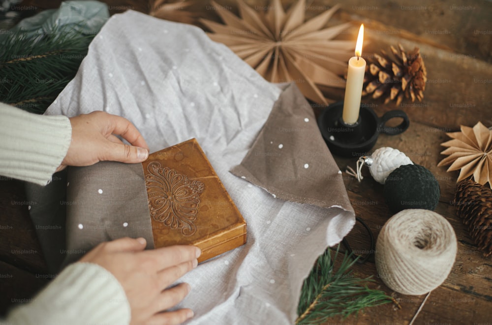 Hands wrapping christmas gift box in brown fabric on rustic wooden table with scissors, craft paper star, candle. Zero waste and eco friendly presents. Atmospheric moody time, nordic style.
