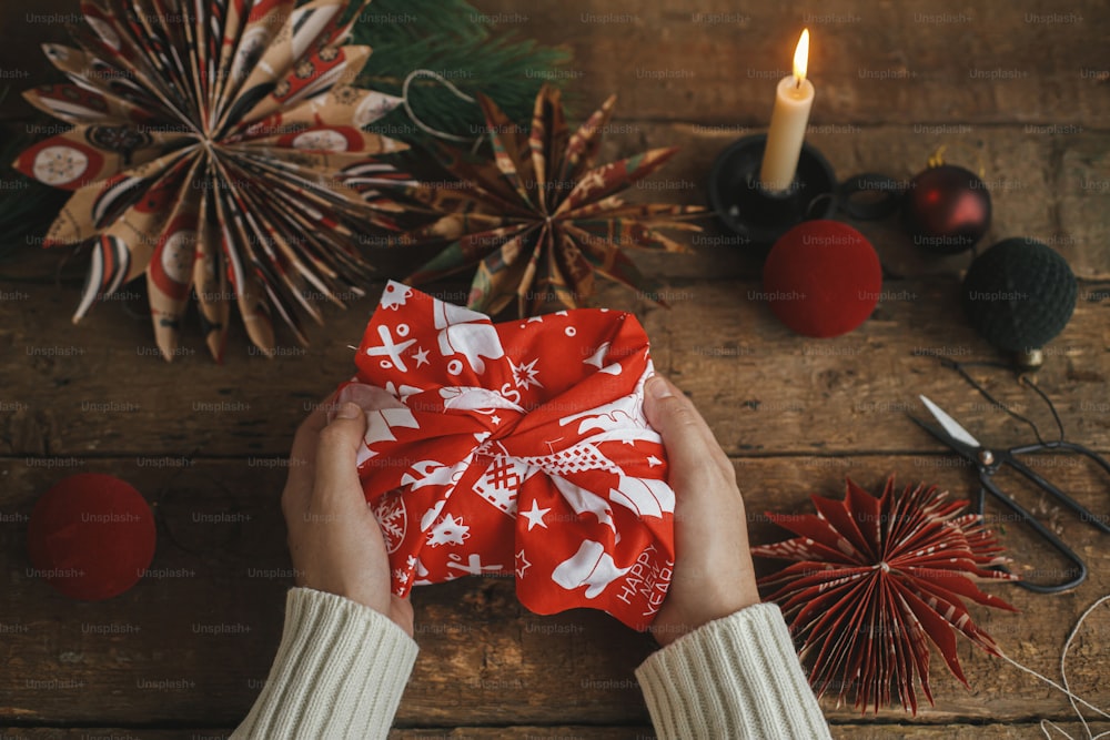 Furoshiki christmas gift. Hands holding christmas gift wrapped in red festive fabric on rustic wooden table with scissors, paper star, candle, ornaments. Atmospheric moody time. Zero waste holiday