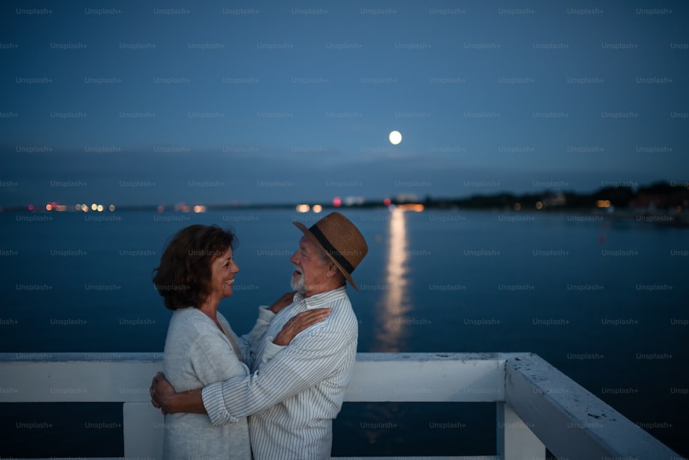 Ein glückliches verliebtes älteres Paar, das sich draußen auf dem Pier am Meer bei Mondschein umarmt und sich gegenseitig ansieht.