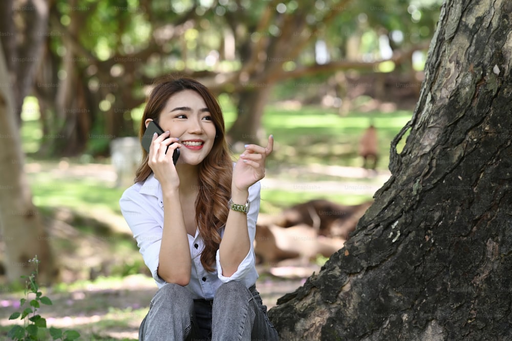 Donna sorridente che si siede nel parco pubblico e parla al telefono cellulare.