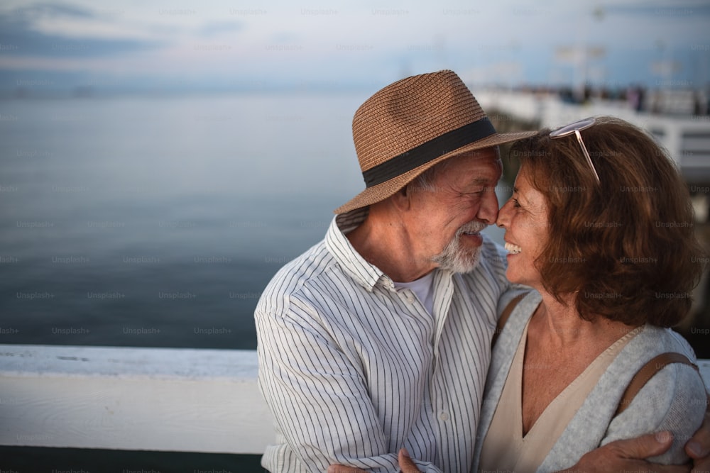 Portrait of happy senior couple in love hugging outdoors on a pier by sea, looking at each other, summer holiday.