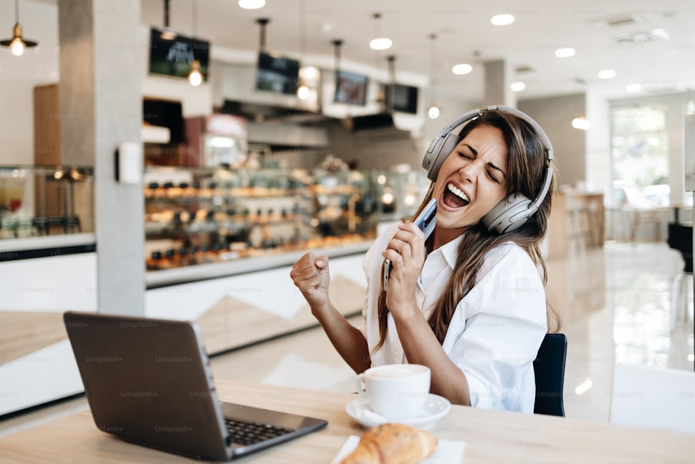 Mujer joven hermosa y feliz sentada y comiendo deliciosos panecillos en panadería o comida rápida. También usa su computadora portátil y escucha música con auriculares.