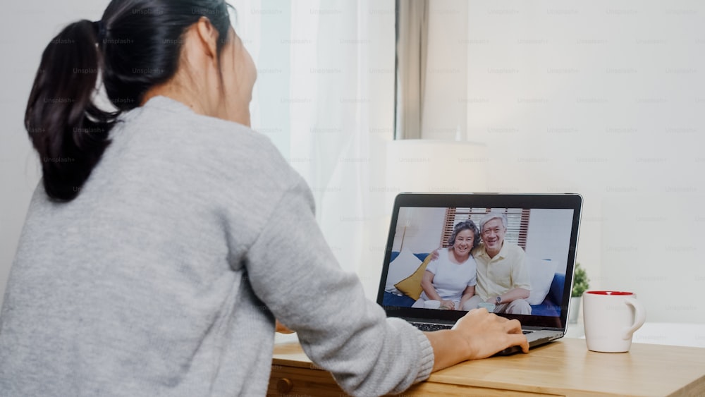 Giovane donna d'affari asiatica che utilizza la videochiamata del laptop che parla con il papà e la mamma di famiglia mentre lavora da casa nella stanza da letto. Autoisolamento, distanziamento sociale, quarantena per la prevenzione del coronavirus.