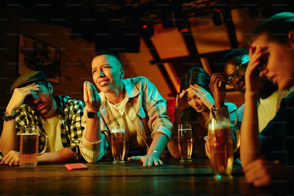 Gruppo multietnico di tifosi scontenti che guardano una partita sportiva in TV in un bar.