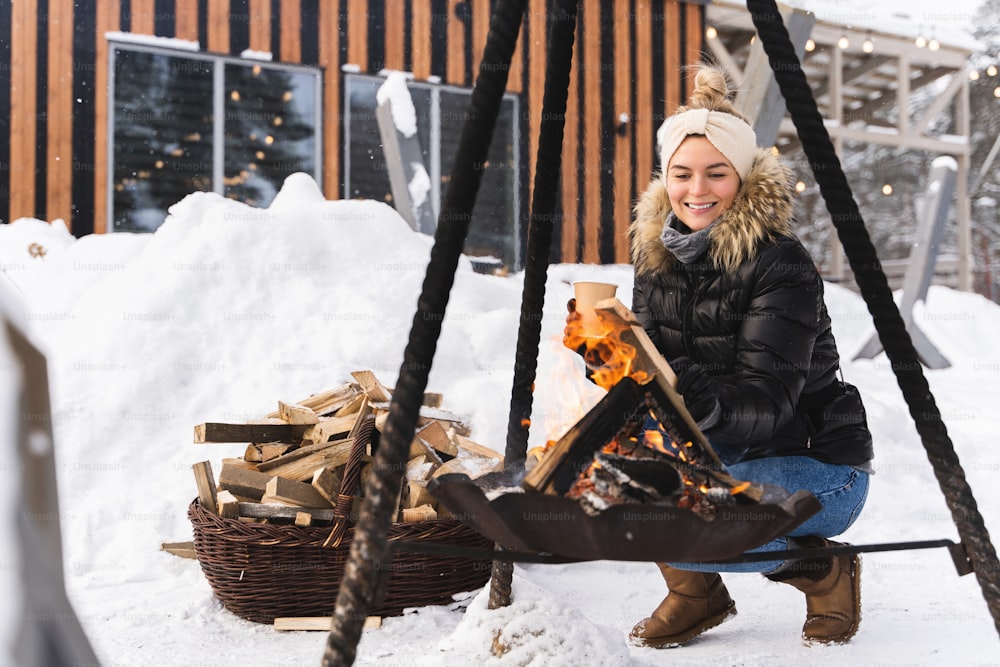 Young and beautiful woman wearing down jacket warming up by the fire pit during cold winter day