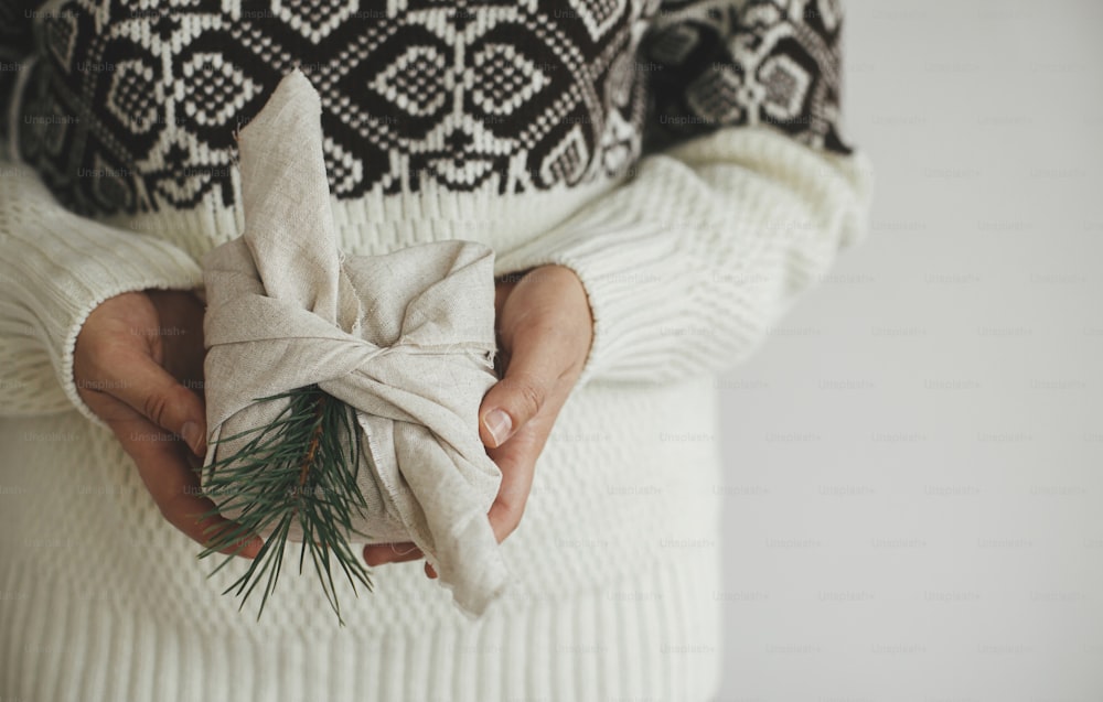Frau Hände in kuscheligen Pullover mit Weihnachtsgeschenk in Stoff verpackt in skandinavischen Raum. Atmosphärisches stimmungsvolles Bild, nordischer Stil. Zero Waste und umweltfreundliche Geschenke, Weihnachtsgeschenk Furoshiki