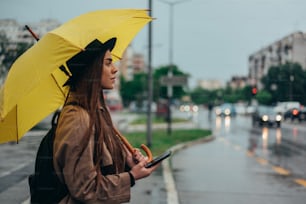 Hermosa mujer joven que usa un teléfono inteligente y sostiene un paraguas amarillo mientras está en la ciudad mientras llueve