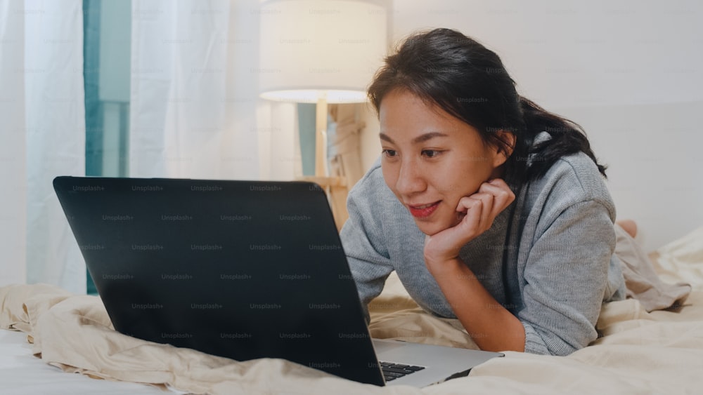 Portrait of freelance Asia woman casual wear using laptop on bed in bedroom at house night. Working from home, remotely work, self isolation, social distancing, quarantine for corona virus prevention.