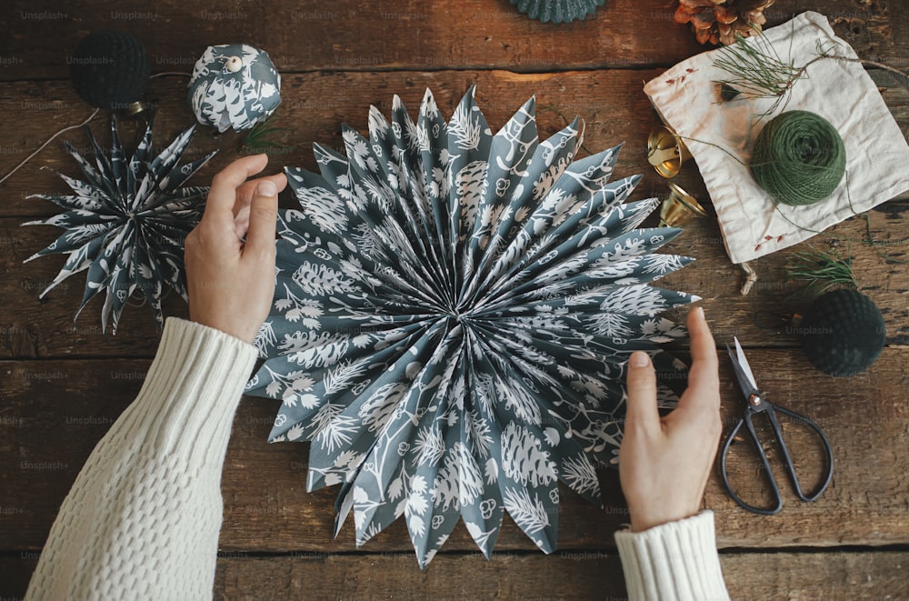 Hände mit stilvollem Weihnachtsstern aus blauem Geschenkpapier auf rustikalem Holztisch flach gelegt. Atmosphärisches stimmungsvolles Bild, nordischer Stil. Handgemachte festliche Dekoration, Urlaubsvorbereitung