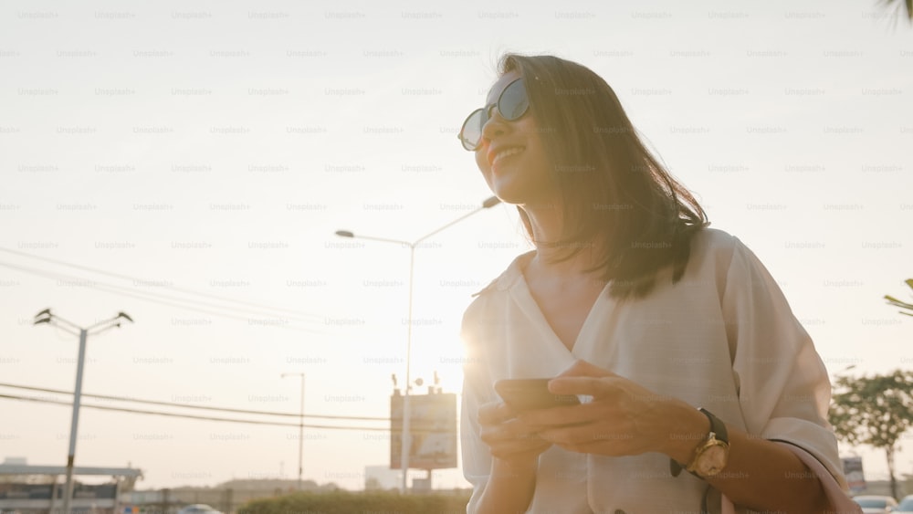 Erfolgreiche junge asiatische Geschäftsfrau in Mode-Bürokleidung, die auf der Straße ein Taxi nimmt und ihr Smartphone hält, während sie draußen in einer urbanen modernen Stadt steht. Business on the go-Konzept.