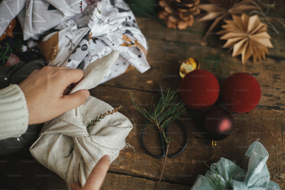 Zero Waste und umweltfreundliche Geschenke, Weihnachts-Furoshiki-Geschenk. Hände wickeln Weihnachtsgeschenk in Stoff auf rustikalem Holztisch mit Ornamenten. Atmosphärisches stimmungsvolles Bild, nordischer Stil.