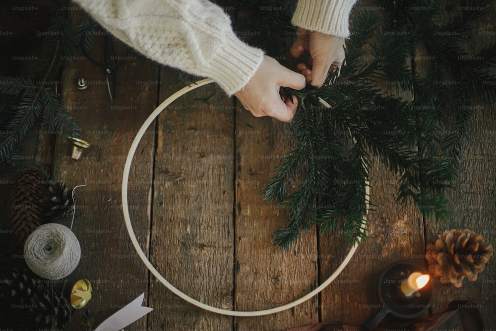 Hands making modern christmas wreath with fir branches on rustic table with round wooden hoop, scissors, thread, candle, pine cones. top view. Atmospheric moody image. Winter holidays preparation