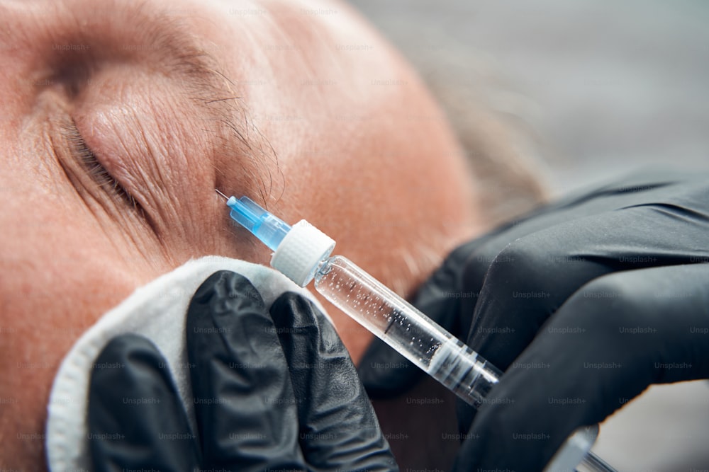 Close up of doctor cosmetologist in sterile gloves using syringe and cotton pad while making injection in man face