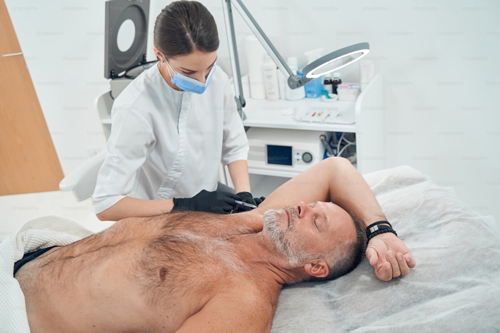 Médecin cosmétologue dans un masque médical faisant une injection dans l’aisselle masculine pendant que l’homme est allongé sur un lit de repos et garde les yeux fermés