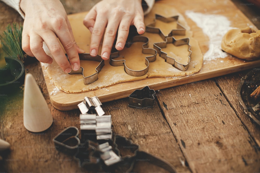 Hand cutting gingerbread dough with christmas metal cutters on wooden board, moody image. Making traditional christmas gingerbread cookies on rustic table with spices, decorations, rolling pin