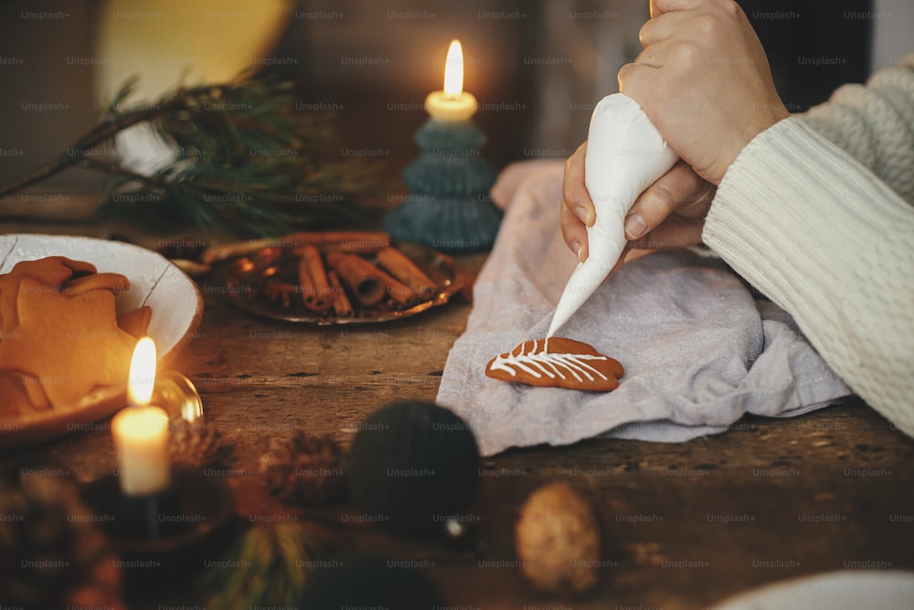 Hände schmücken Lebkuchenplätzchen Weihnachtsbaum mit Zuckerguss auf rustikalem Tisch mit Serviette, Kerze, Gewürzen, Dekorationen. Atmosphärisches stimmungsvolles Bild. Herstellung traditioneller Weihnachtslebkuchenplätzchen