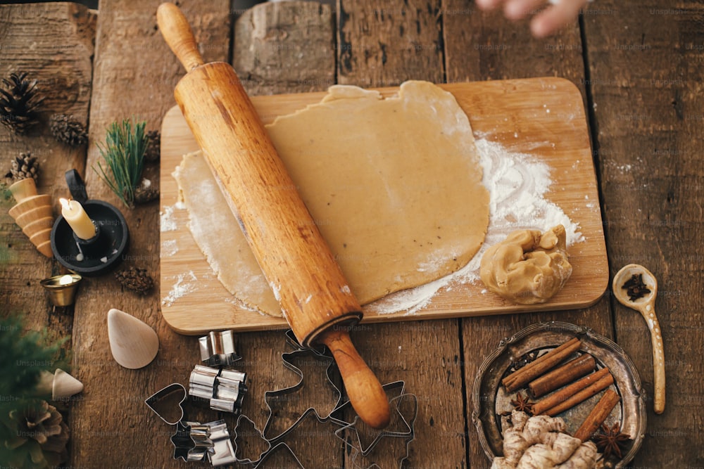 Gingerbread dough with wooden rolling pin on rustic wooden table with candle, metal cutters, spices, decorations. Making traditional christmas gingerbread cookies, rustic flat lay