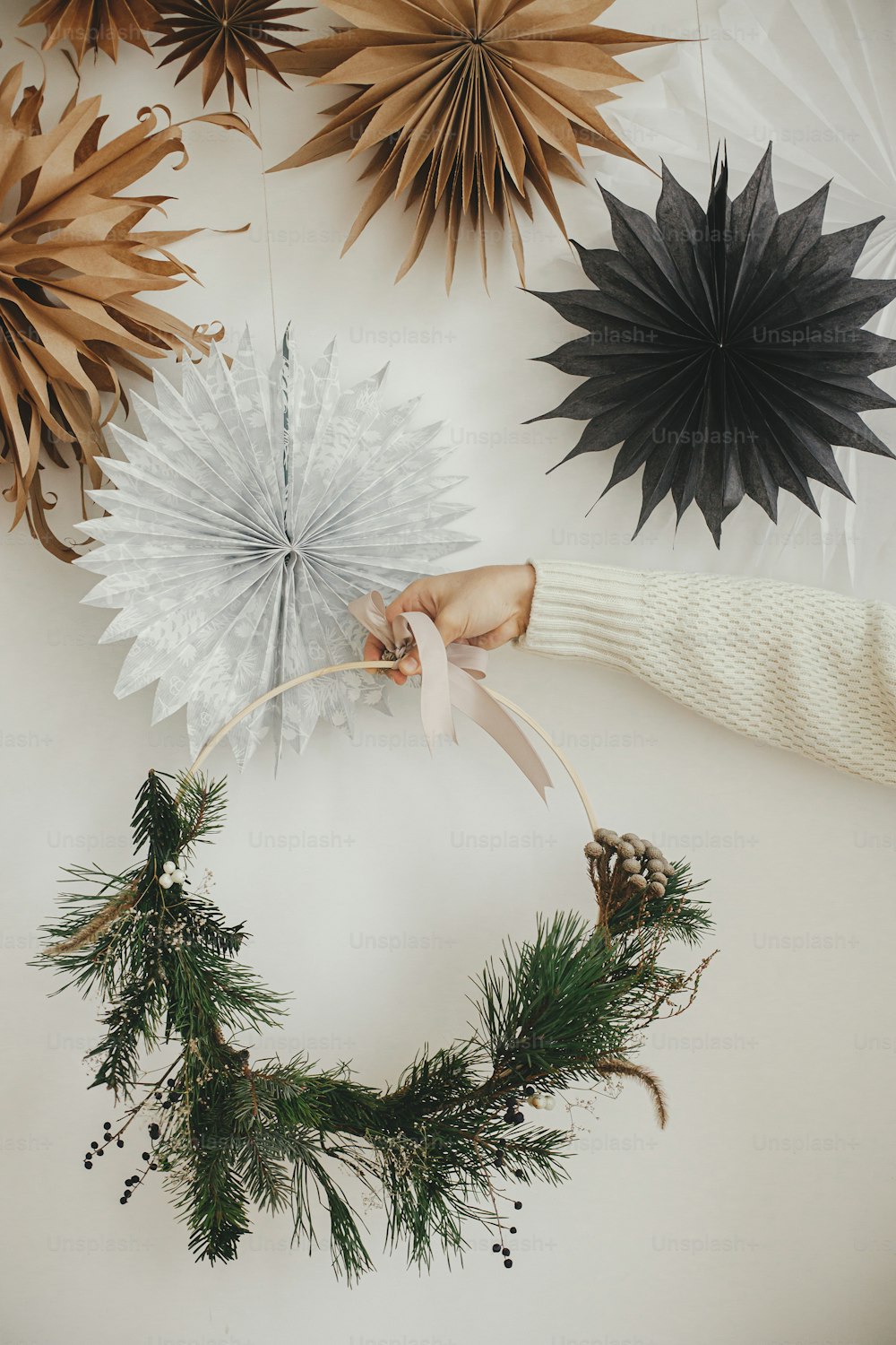 Hand in cozy sweater holding modern christmas wreath on white wall background with swedish paper stars. Merry Christmas and Happy holidays! Minimalist xmas boho wreath in hands. Atmospheric time