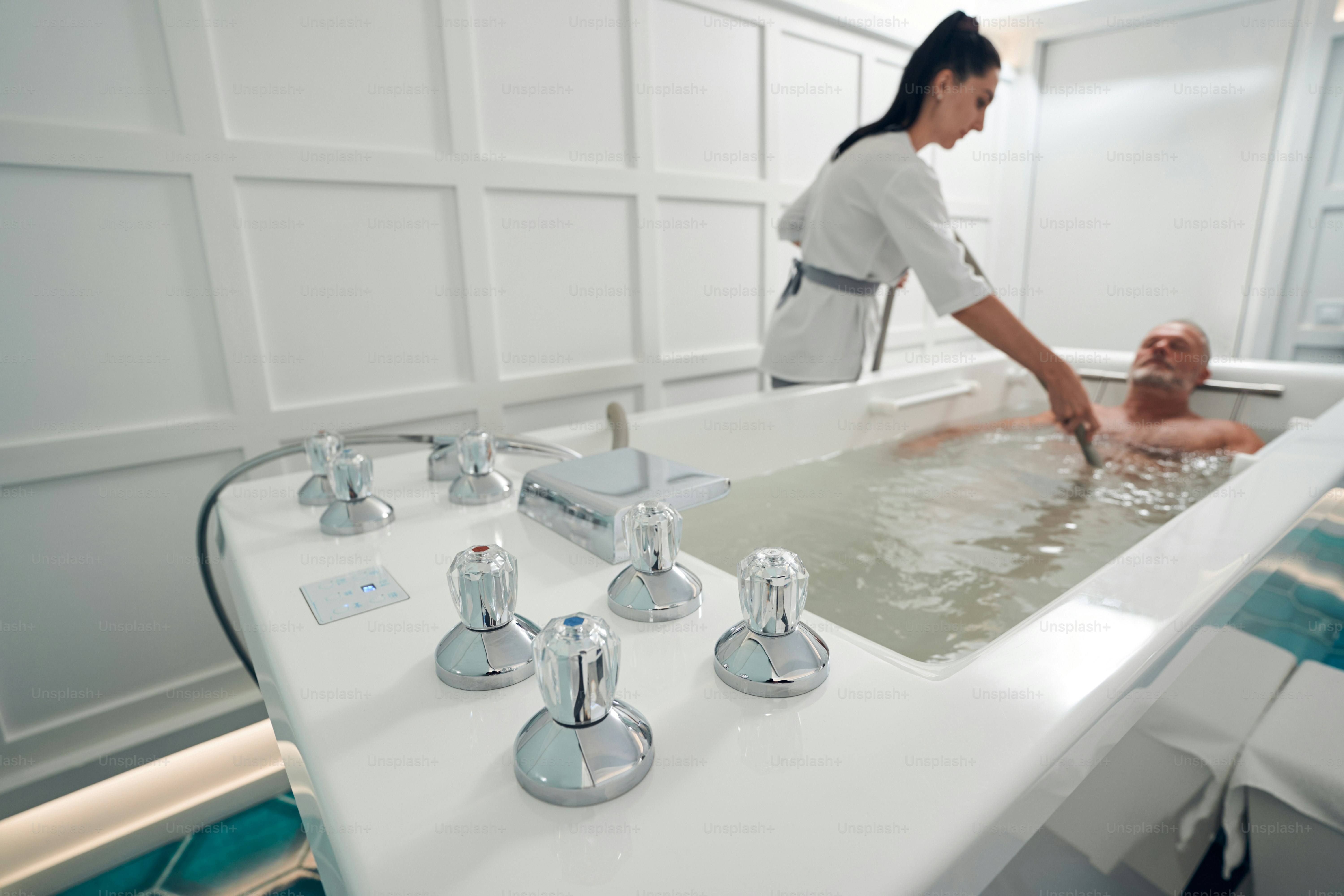 Male person lying in bathtub with metal thermostatic parts while beautician performing hydromassage with special device