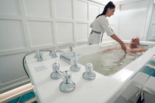 Persona masculina acostada en bañera con partes termostáticas de metal mientras esteticista realiza hidromasaje con dispositivo especial