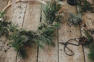 Corona navideña moderna con ramas de abeto, hierbas, tijeras, piñas sobre madera rústica. ¡Feliz Navidad! Elegantes detalles de corona navideña boho. Imagen malhumorada, preparación para las vacaciones de invierno