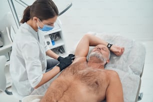 Doctor in medical mask making injection in male armpit while man lying on daybed and keeping eyes closed