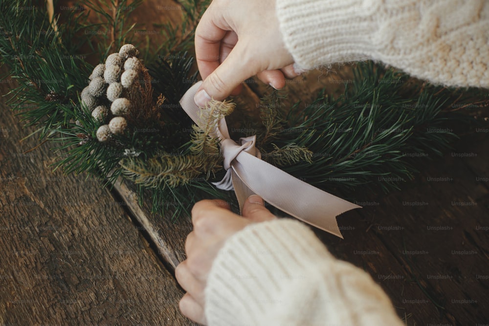 Hands tying ribbon and making modern christmas wreath on rustic table with fir branches, brunia herb, round wooden hoop. Atmospheric moody image. Winter holidays preparation close up