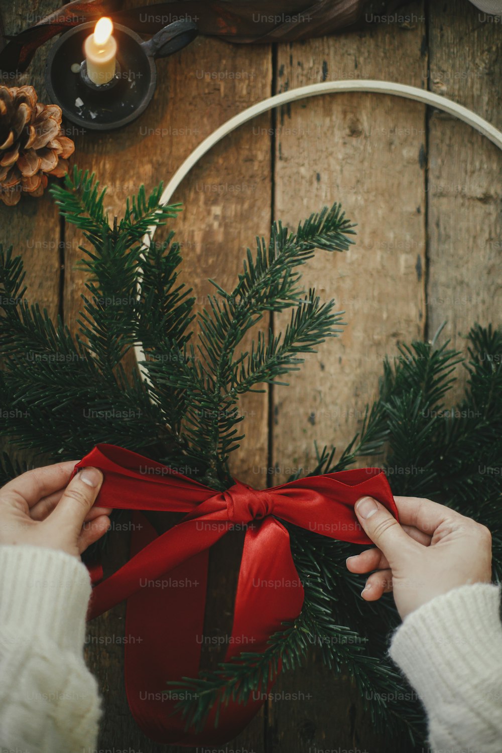 Mãos fazendo coroa de Natal moderna com ramos de abeto e fita vermelha na mesa rústica com vela, fitas, pinhas. Vista superior.  Imagem atmosférica mal-humorada. Preparação para as férias de inverno