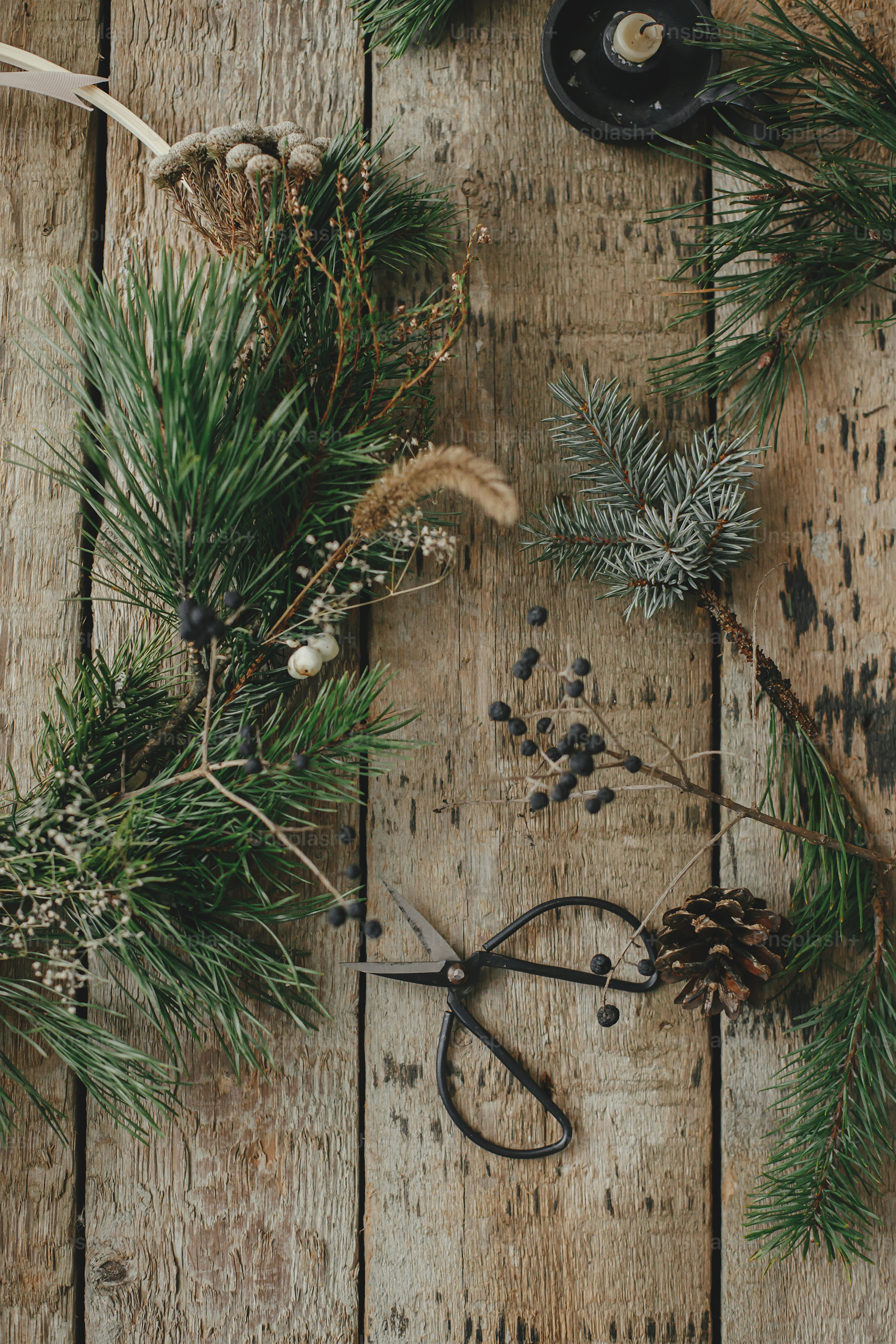 Modern christmas wreath flat lay. Stylish xmas wreath with spruce branches, herbs, scissors,candle, pine cones on rustic wood. Moody image. Merry Christmas! Winter holidays preparation