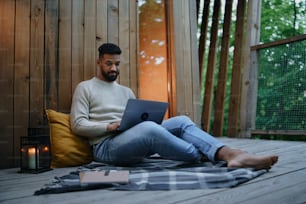 Um jovem feliz com laptop descansando ao ar livre em uma casa na árvore, fim de semana e conceito de escritório remoto.