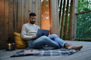 A happy young man with laptop resting outdoors in a tree house, weekend away and remote office concept.