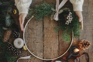 Hands in sweater holding modern christmas wreath on rustic wooden background flat lay. Boho wreath on rustic table with thread, candle, pine cones. Atmospheric image. Winter holiday preparation