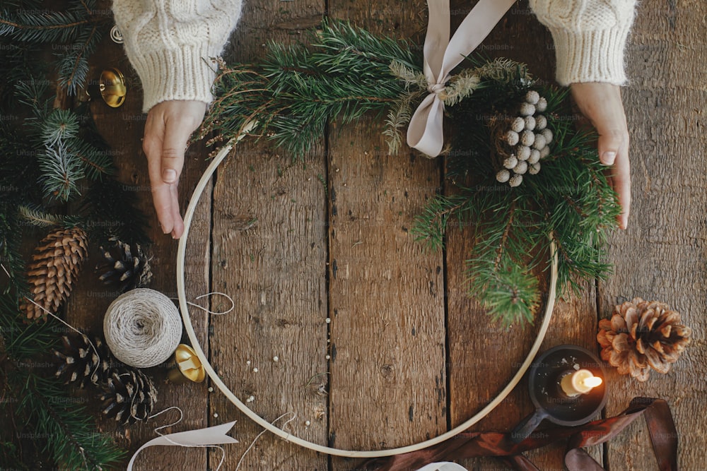 Hands in sweater holding modern christmas wreath on rustic wooden background flat lay. Boho wreath on rustic table with thread, candle, pine cones. Atmospheric image. Winter holiday preparation