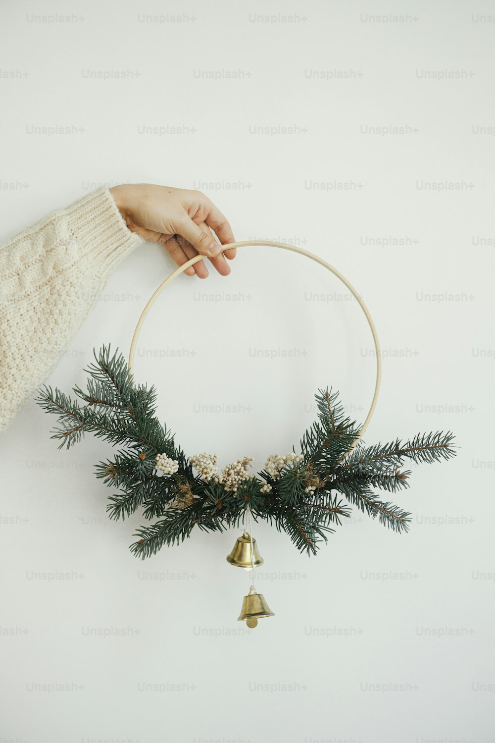 Hand in cozy sweater holding modern christmas wreath on white wall background. Merry Christmas and Happy holidays! Minimalist xmas boho wreath with bells in hand. Atmospheric time