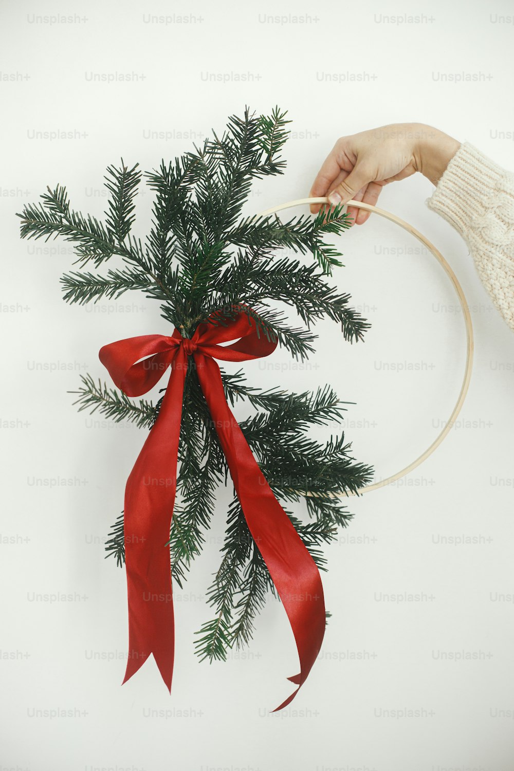 Hand in cozy sweater holding modern christmas wreath on white wall background. Merry Christmas and Happy holidays! Stylish minimalistic xmas wreath with fir branches and red bow in hand