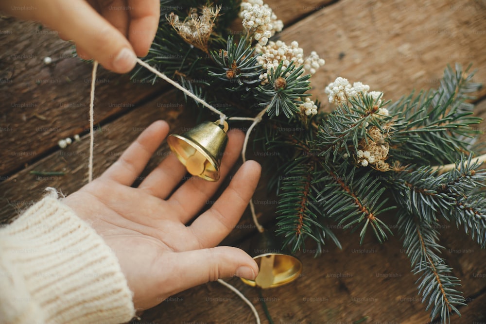 Making modern christmas wreath. Hands decorating xmas boho wreath with bells, fir branches, herbs on rustic table. Atmospheric moody image. Winter holidays preparation. Merry Christmas!