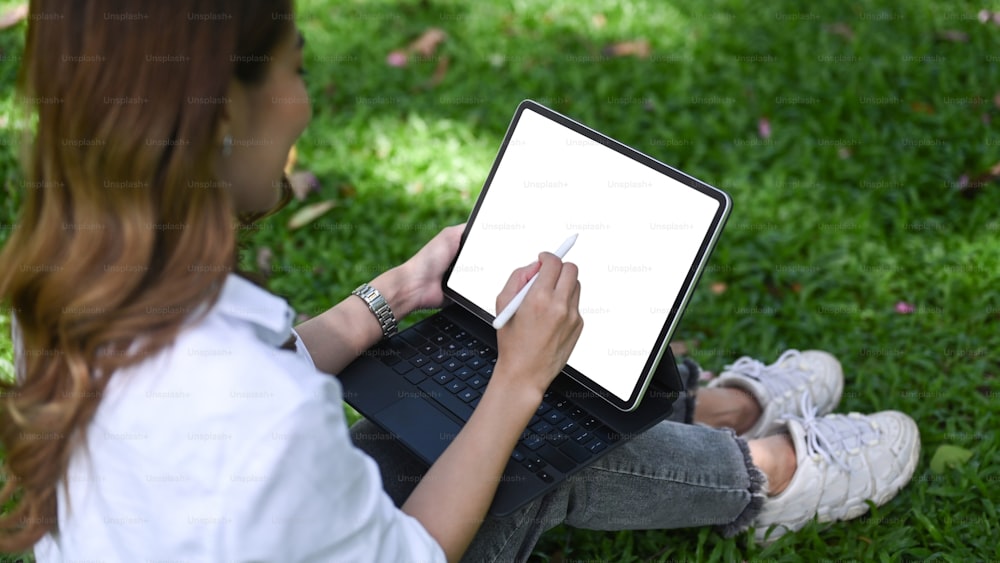 웃고 있는 여성 프리랜서는 공원의 잔디밭에 앉아 디지털 태블릿으로 작업하고 있다.