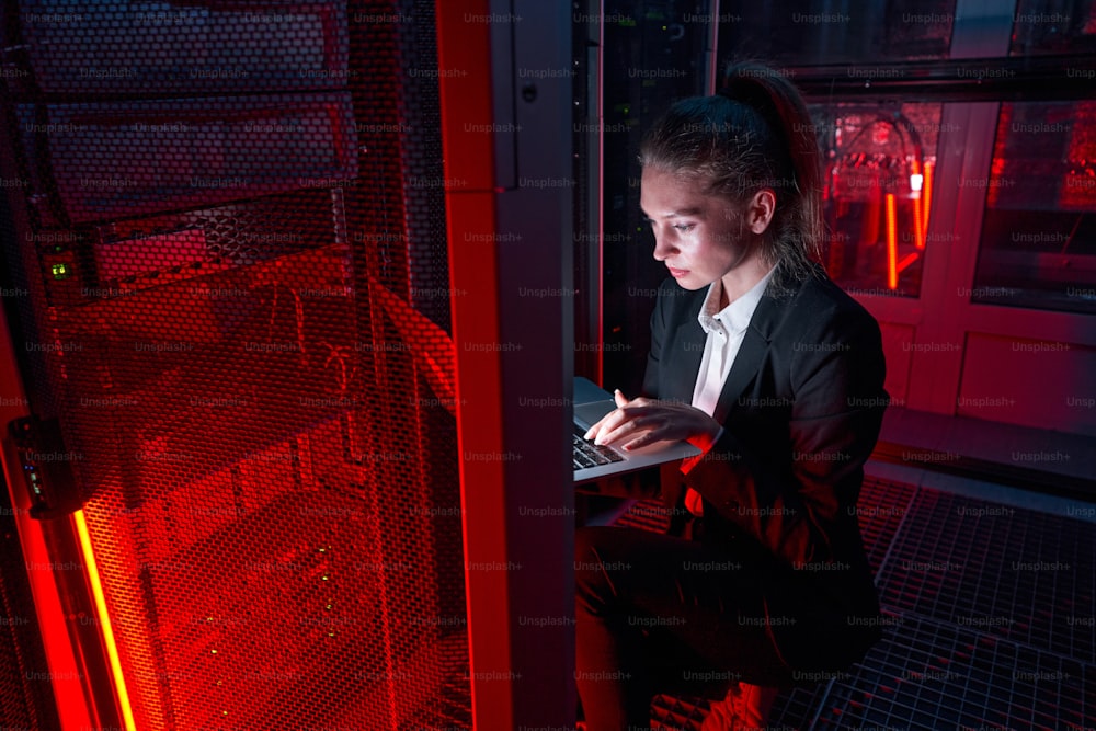 Ernsthafte kaukasische Ingenieurin, die neben Servergeräten hockt und einen Laptop benutzt, während sie das Problem im von rotem Licht beleuchteten Serverraum behebt