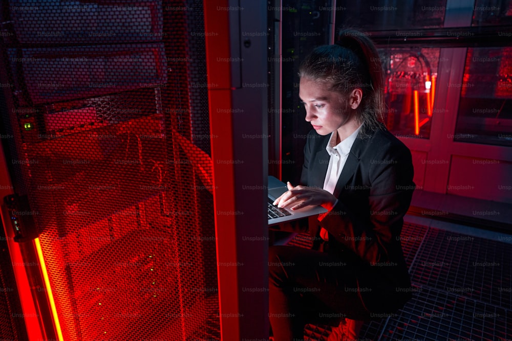 Une ingénieure caucasienne sérieuse accroupie près de l’équipement du serveur et utilisant un ordinateur portable tout en dépannant un problème dans la salle des serveurs éclairée par une lumière rouge