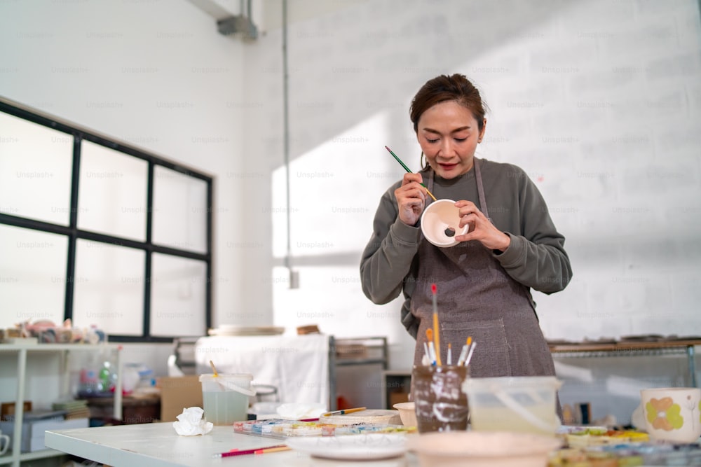 Asian woman learning color painting her self-made pottery at home. Confidence female enjoy hobbies and indoors leisure activity handicraft ceramic sculpture and painting workshop at pottery studio.