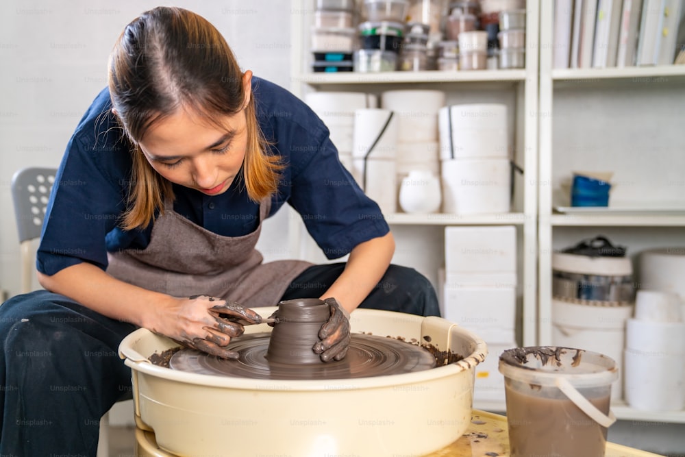 아름 다운 아시아 여성 조각가 예술가는 세라믹 스튜디오에서 도자기 바퀴에 점토를 조각합니다. 생 점토를 성형하는 여성 장인은 작업장에서 도자기 모양을 만듭니다. 중소 기업 수공예 제품 개념입니다.
