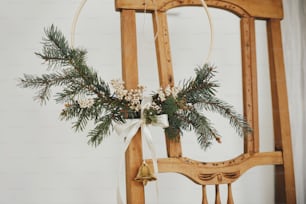 Modern christmas wreath with bell hanging on rustic wooden chair. Winter holidays preparation, atmospheric moody image. Stylish boho xmas wreath in scandinavian room. Merry Christmas!