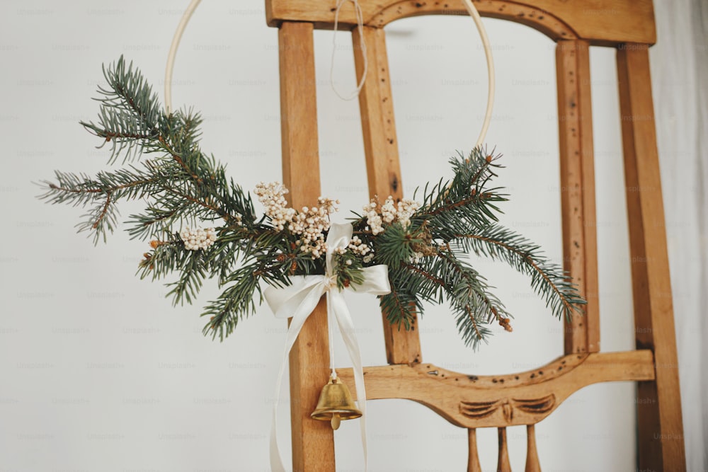 Moderner Weihnachtskranz mit Glocke auf rustikalem Holzstuhl. Winterurlaubsvorbereitung, stimmungsvolles Stimmungsbild. Stilvoller Boho-Weihnachtskranz im skandinavischen Zimmer. Frohe Weihnachten!