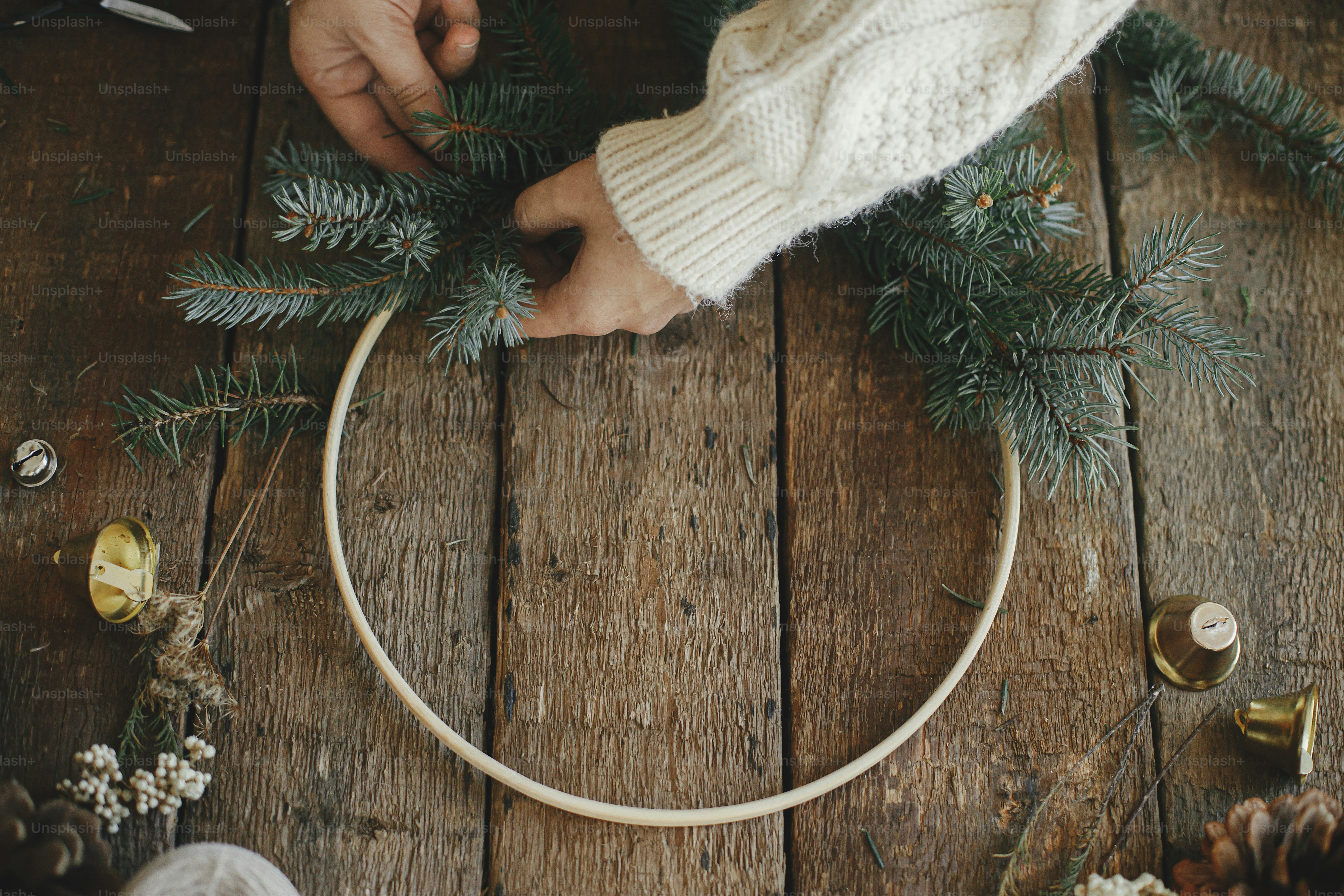 Hands in cozy sweater making modern christmas wreath with fir branches, herbs, bells, round wooden hoop on rustic table. Atmospheric moody image. Making xmas boho wreath